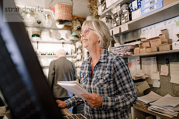 Lächelnde ältere Verkäuferin  die wegschaut  während sie mit Dokumenten an der Kasse im Laden steht