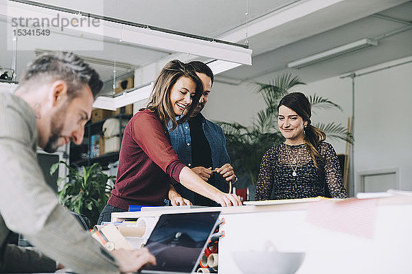 Lächelnde Geschäftsfrau arbeitet mit Kollegen zusammen  während ein Geschäftsmann seinen Laptop im Kreativbüro benutzt