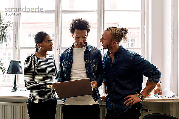 Männlicher Unternehmer hält Laptop in der Hand  während er mit Kollegen gegen Fenster im Büro diskutiert