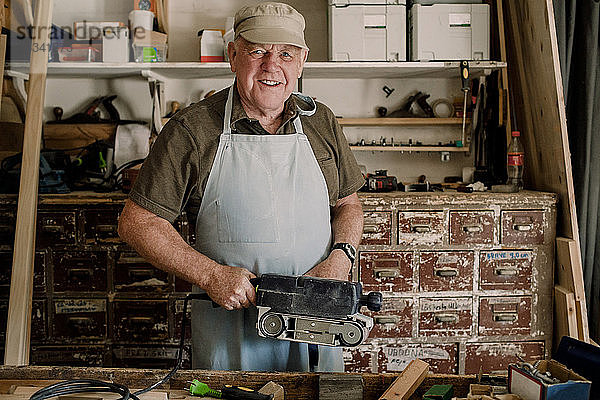 Porträt eines selbstbewusst lächelnden älteren männlichen Eigentümers  der bei einem Workshop ein Elektrowerkzeug hält