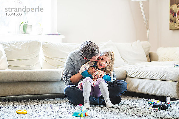 Mittelgroßer Erwachsener Mann küsst Tochter  während er zu Hause auf einem Teppich sitzt