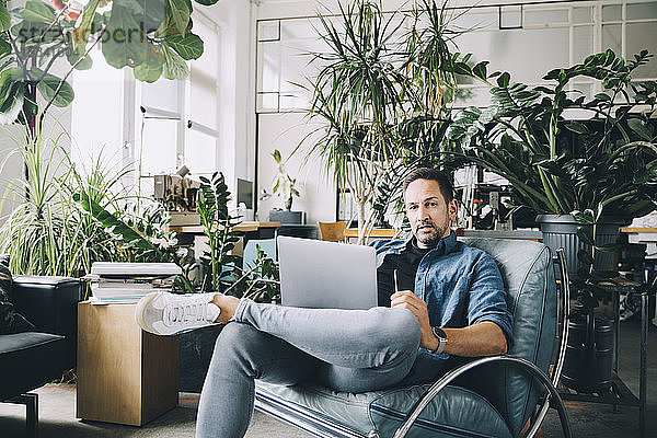 Porträt eines selbstbewussten  kreativen Geschäftsmannes mit Mobiltelefon und Laptop auf einem Sitz im Büro