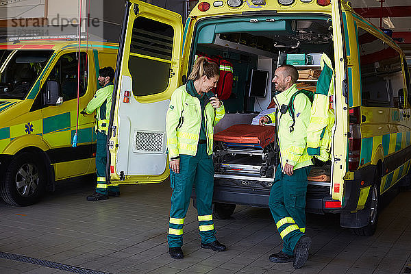 Lächelnder männlicher Sanitäter  der eine Mitarbeiterin ansieht  die mit einem Walkie-Talkie spricht  während er auf dem Parkplatz beim Krankenwagen steht