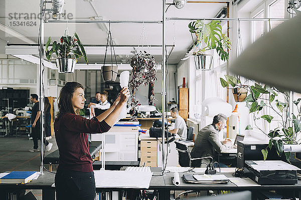 Mittlere erwachsene Geschäftsfrau prüft Papier  während Kollegen im Hintergrund im Kreativbüro arbeiten
