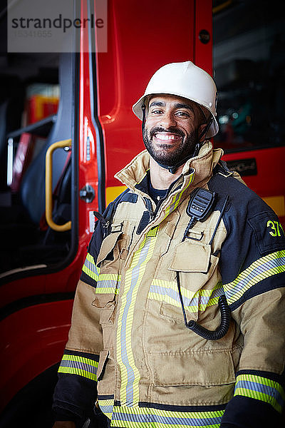 Porträt eines lächelnden männlichen Feuerwehrmannes  der sich in der Feuerwache gegen ein Feuerwehrauto stellt