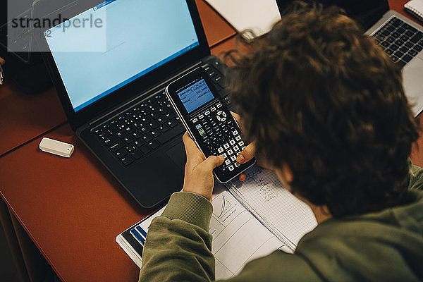 Hochwinkelansicht eines Teenagers  der einen Taschenrechner benutzt  während er über einen Laptop am Schreibtisch im Klassenzimmer lernt