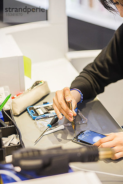 Schrägaufnahme einer jungen Arbeitnehmerin bei der Reparatur eines beschädigten Smartphones am beleuchteten Schreibtisch in einer mobilen Reparaturwerkstatt