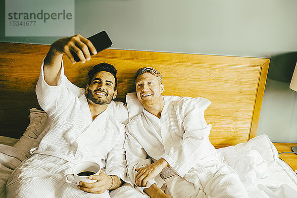 Lächelnder Schwuler mit Kaffeetasse in der Hand  während er mit seinem Freund im Hotelzimmer Selfie nimmt