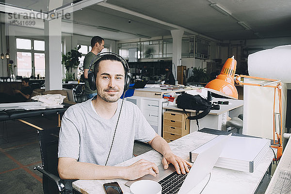 Porträt eines selbstbewussten Geschäftsmannes mit Laptop  während ein männlicher Kollege im Hintergrund im Kreativbüro arbeitet