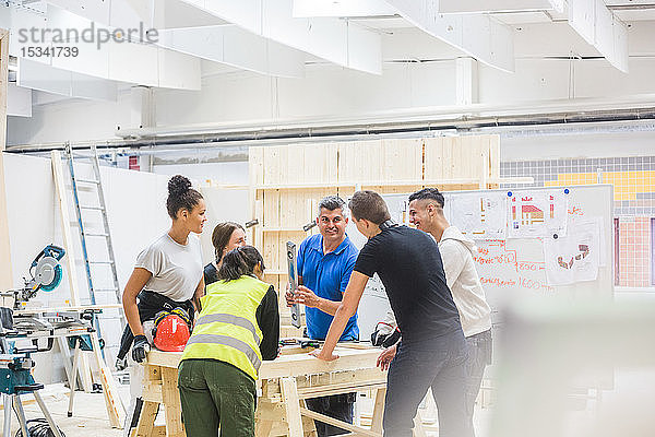 Männlicher Ausbilder unterrichtet Auszubildende  die an der Werkbank in einer beleuchteten Werkstatt stehen  über das Niveau