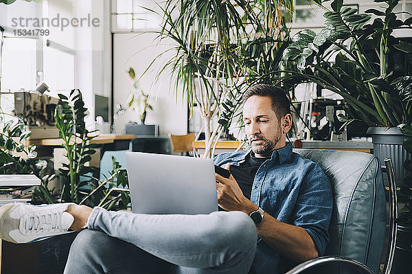 Selbstbewusster kreativer Geschäftsmann  der ein Mobiltelefon benutzt  während er im Büro mit einem Laptop auf einem Sitzplatz sitzt