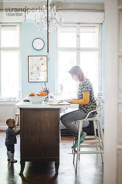 Frau in voller Länge am Laptop arbeitend  während der Sohn zu Hause an der Kücheninsel steht