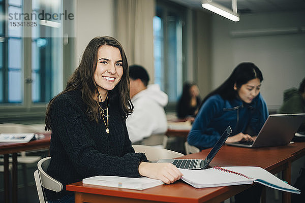 Porträt einer lächelnden Teenagerin am Schreibtisch sitzend mit Klassenkameraden im Hintergrund