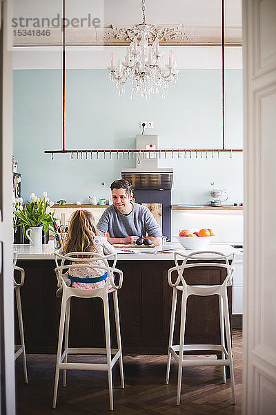 Lächelnder Vater sieht Mädchen beim Lernen an  während er an einer Kücheninsel sitzt