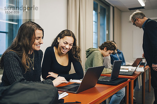 Fröhliche Gymnasiastinnen mit Laptop am Schreibtisch im Klassenzimmer