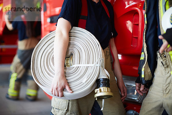 Mittlere Sektion einer weiblichen Feuerwehrfrau  die einen aufgerollten Feuerwehrschlauch hält  während sie mit einem Kollegen in der Feuerwache steht