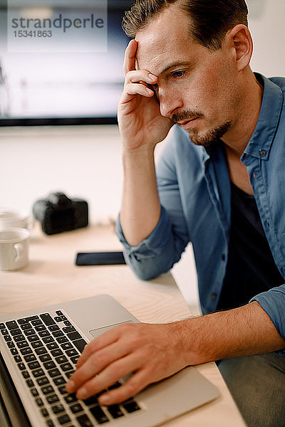 Besorgter Geschäftsmann benutzt Laptop  während er im Kreativbüro sitzt
