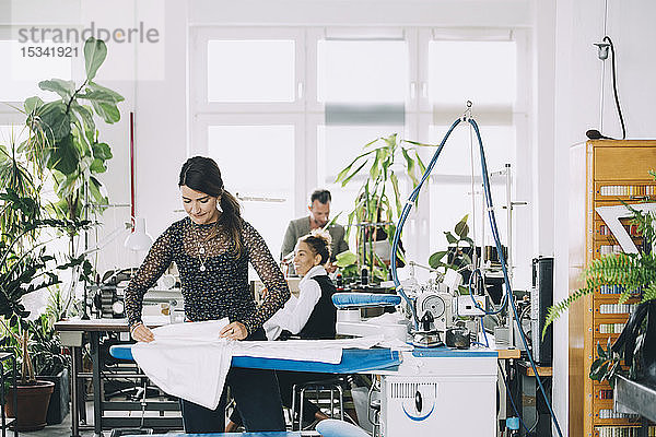 Unternehmerin bügelt weißen Stoff  während Kollegen im Hintergrund im Kreativbüro arbeiten
