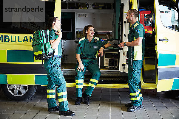 Weibliche Rettungssanitäterin sitzt im Krankenwagen  während sie sich auf dem Parkplatz mit Kollegen unterhält
