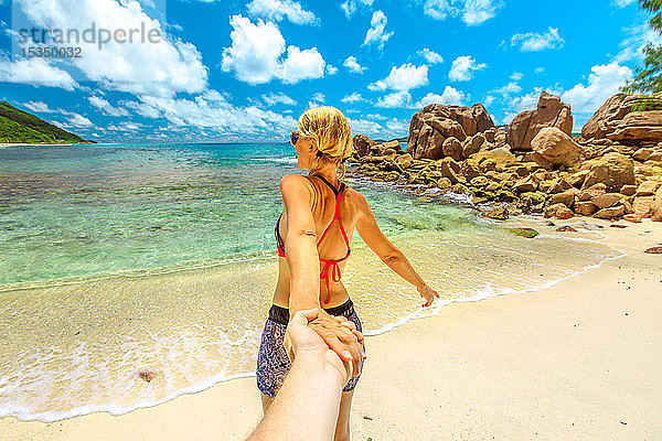 Blonde Touristin im Bikini hält die Hand ihres Partners am Anse Caiman Beach mit natürlichem Pool in La Digue  Seychellen  Indischer Ozean  Afrika