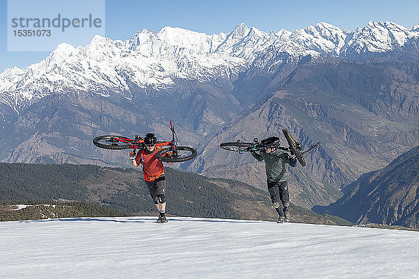 Mountainbiker tragen ihre Fahrräder einen schneebedeckten Hang im Himalaya hinauf  mit Blick auf die Langtang-Kette in der Ferne  Nepal  Asien