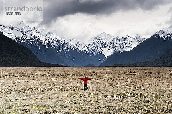 Mann in rotem Mantel steht mit ausgebreiteten Armen und schaut auf schneebedeckte Berge  Fiordland National Park  UNESCO Weltkulturerbe  Südinsel  Neuseeland  Pazifik