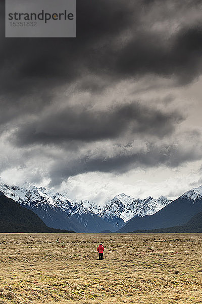 Mann in rotem Mantel mit Blick auf schneebedeckte Berge  Fiordland National Park  UNESCO Weltkulturerbe  Südinsel  Neuseeland  Pazifik