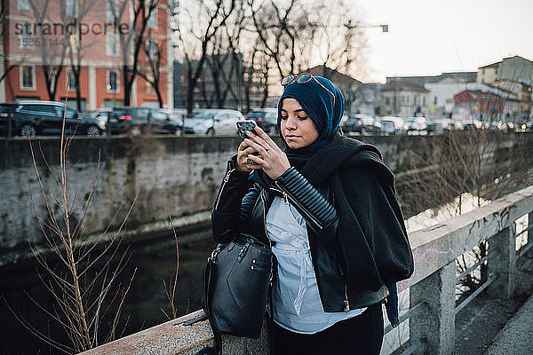 Junge Frau im Hidschab betrachtet Smartphone am Kanal in der Stadt