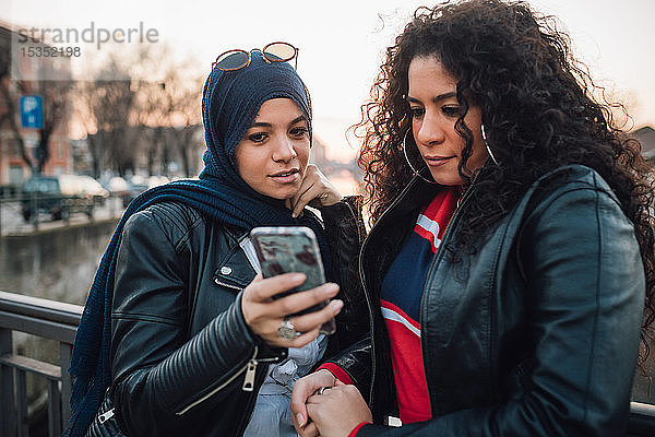 Junge Frau im Hidschab und beste Freundin schauen sich Smartphone auf der Kanalbrücke in der Stadt an