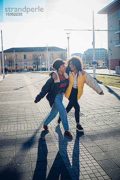 Zwei coole junge Freundinnen schlendern und lachen auf dem städtischen Bürgersteig