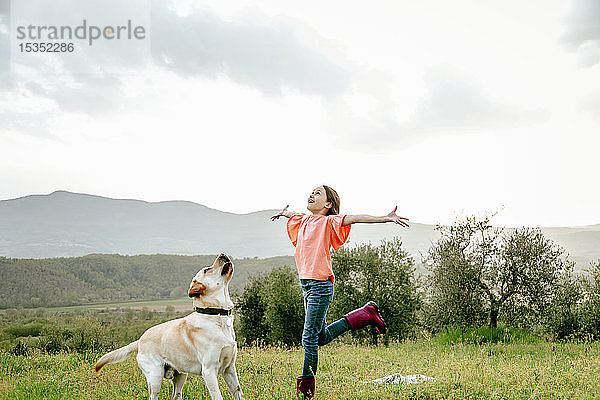 Mädchen mit offenen Armen spielt mit Labradorhund in malerischer Feldlandschaft  Citta della Pieve  Umbrien  Italien