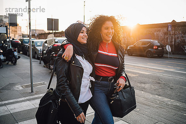Junge Frau im Hidschab und beste Freundin gehen und reden bei Sonnenuntergang in der Stadt
