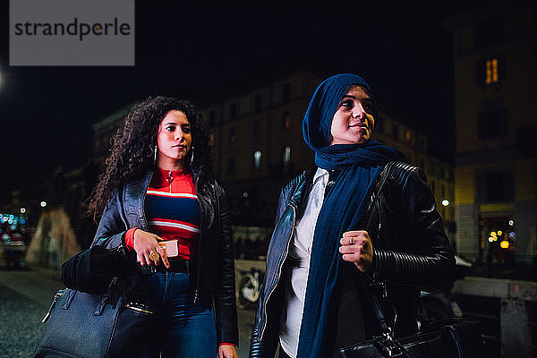 Junge Frau im Hidschab und beste Freundin nachts auf dem Bürgersteig der Stadt