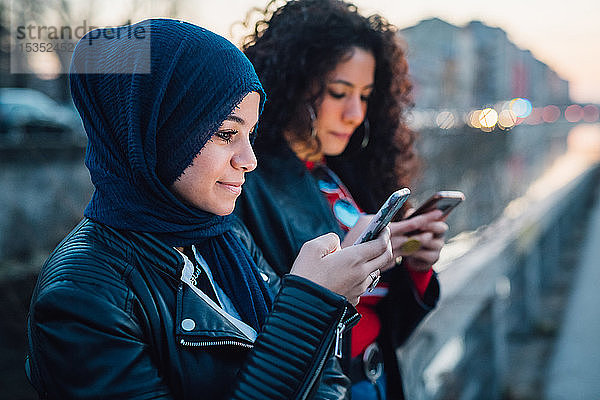 Junge Frau im Hidschab mit bester Freundin beim Betrachten von Smartphones am Stadtkanal