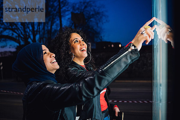 Junge Frau im Hijab mit bester Freundin mit beleuchtetem Touchscreen an nächtlicher Bushaltestelle