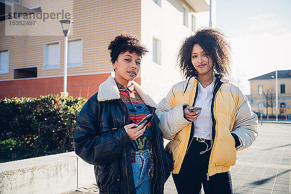 Zwei coole junge Freundinnen mit Smartphones auf dem städtischen Bürgersteig  Porträt