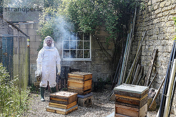 Männlicher Imker trägt Bienenraucher im ummauerten Garten  Porträt