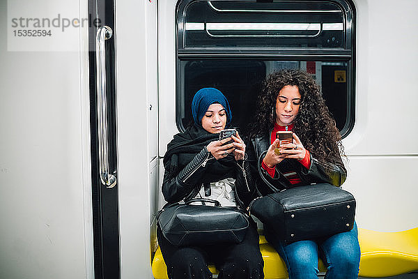 Junge Frau im Hidschab und Freundin sitzen in der U-Bahn und schauen sich Smartphones an