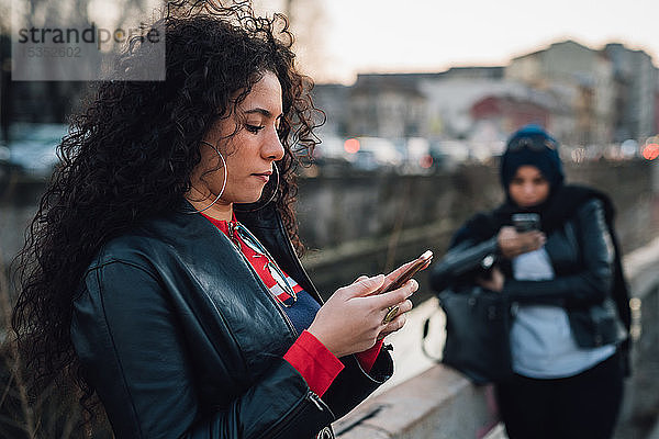 Mittelgroße erwachsene Frau mit langen lockigen Haaren betrachtet Smartphone am Stadtkanal