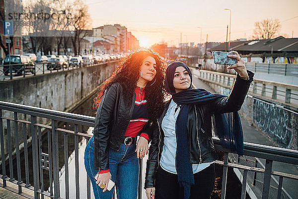 Junge Frau im Hidschab und beste Freundin bei Sonnenuntergang auf der Kanalbrücke in der Stadt