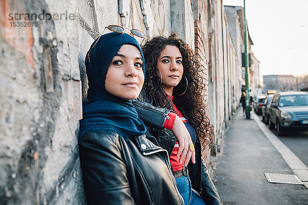 Junge Frau im Hidschab und beste Freundin lehnen an der Wand auf dem Bürgersteig der Stadt
