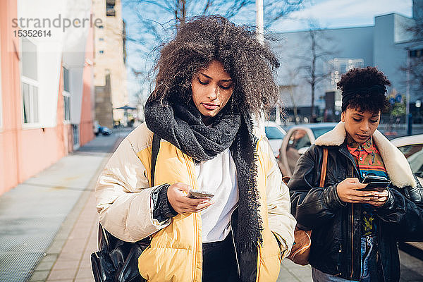 Zwei coole junge Frauen  die auf dem städtischen Bürgersteig spazieren gehen und sich Smartphones anschauen