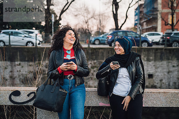 Junge Frau im Hidschab und ihr Freund benutzen Smartphones und lachen am Stadtkanal