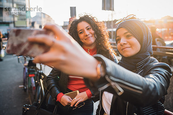 Junge Frau im Hidschab mit Freundin beim Smartphone-Selfie in der Stadt