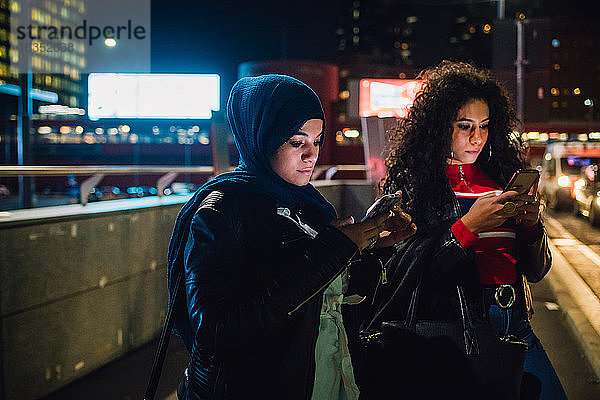 Junge Frau im Hidschab mit Freundin betrachtet nachts Smartphones in der Stadt