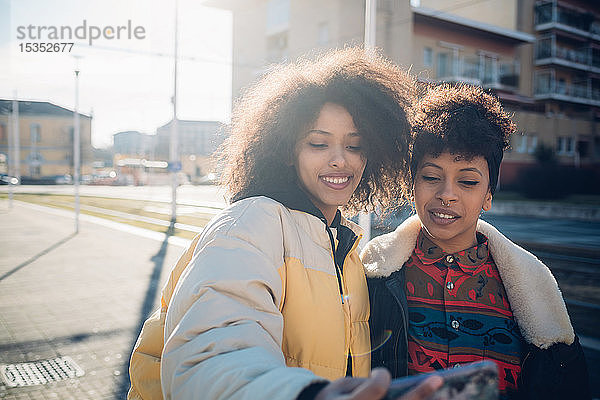 Zwei coole junge Freundinnen mit Smartphone-Selfie auf dem städtischen Bürgersteig