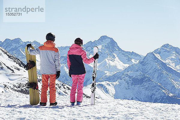 Teenager-Junge Snowboarder mit Schwester  die vom schneebedeckten Berggipfel aus den Blick auf sich zieht  Rückansicht  Alpe-d'Huez  Rhône-Alpes  Frankreich