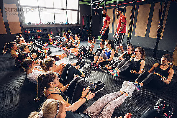 Große Gruppe von Frauen trainiert in Turnhalle mit männlichen Trainern  die in Reihen mit angehobenen Beinen auf dem Boden sitzen