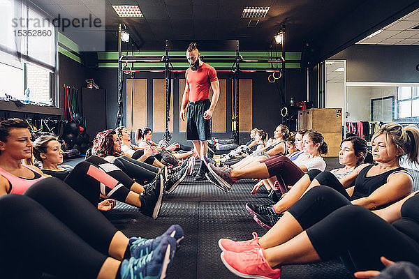 Große Gruppe von Frauen trainiert in Turnhalle mit einem männlichen Trainer  der mit angehobenen Beinen auf dem Boden sitzt