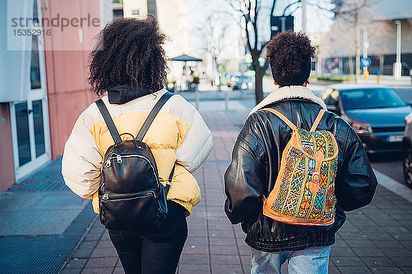 Zwei junge Frauen mit Rucksäcken beim Spaziergang auf dem städtischen Bürgersteig  Rückansicht